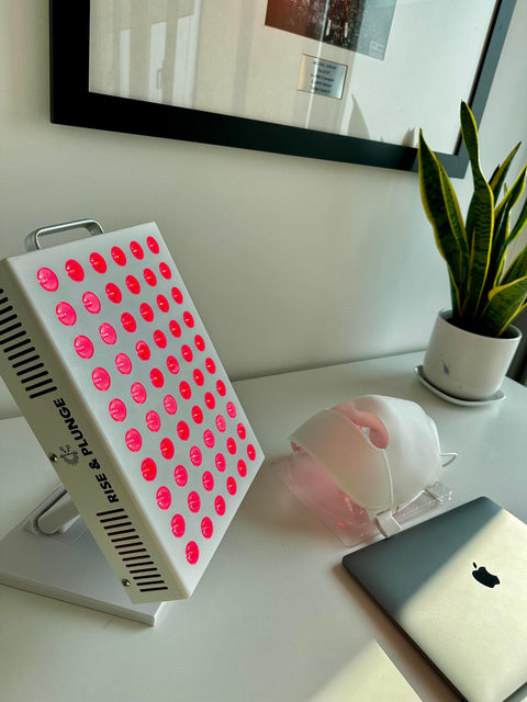Red light therapy panel with pink LEDs on a white desk, near a white chair, a potted plant, and a computer, in a bright office