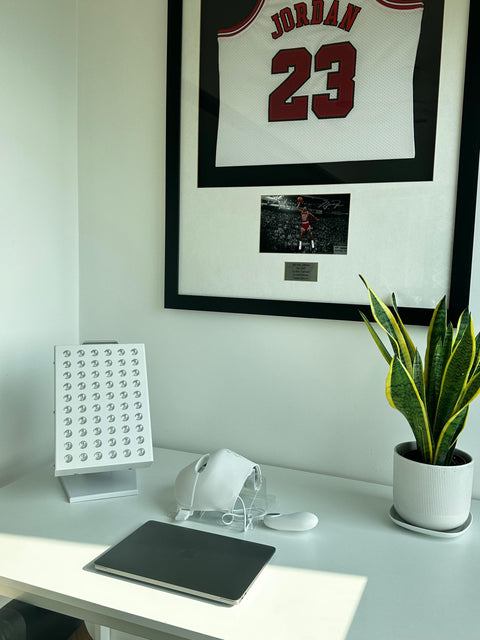 Framed sports jersey and photo of Michael Jordan on a wall, with a white LED red light therapy panel and a plant on a white desk